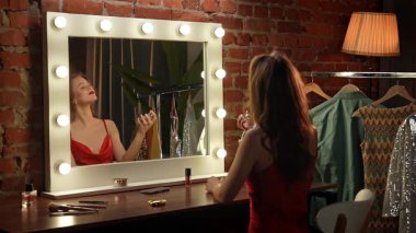 Kırmızı elbiseli bir kadın, şık ışıklandırması ve şık kıyafetleri olan bir soyunma odasına makyaj yapar..