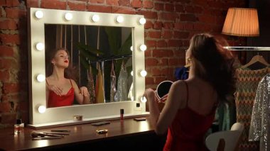 Kırmızı elbiseli bir kadın, şık ışıklandırması ve şık kıyafetleri olan bir soyunma odasına makyaj yapar..