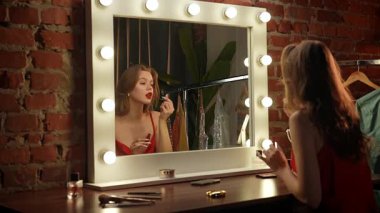 Bir kadın parlak ışıklarla çevrili büyük bir aynanın önünde ruj sürerken görülüyor..