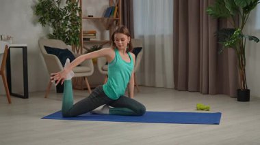 Genç bir kadın, içeride mavi bir minder üzerinde yoga egzersizleri yapıyor, esneklik ve gevşemeye odaklanıyor..