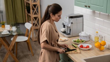 Günlük rutin yaşam tarzı konsepti. Sabahları genç esmer kadın mutfakta sağlıklı kahvaltı hazırlıyor, salatalıkları bıçakla kesiyor, salata yapıyor..
