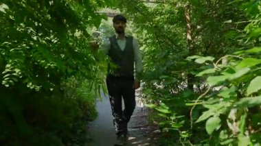 Çelik yelek giyen bir adam yoğun, canlı yeşilliklerle çevrili bir yolda yürür. Huzurlu bir doğal manzara sunar..