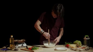 Bir şef, çeşitli sebzeler ve mutfak aletleriyle çevrili bir kasede yemek pişirme işlemini gösteren malzemeleri karıştırır. Yavaş çekim