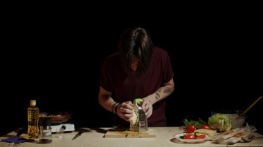 Aşçı bir birey, taze sebzeler ve temel yemek malzemeleriyle çevrili mutfakta rendelenmiş peynirlere odaklanır. Yavaş çekim