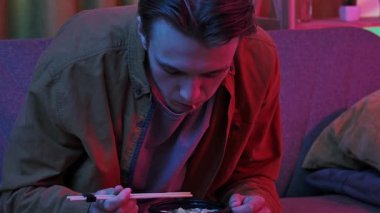 Evde Asya fast food 'unun tadını çıkarıyorum. Genç adam akşam evde kanepede oturuyor, lezzetli Asya yemekleri sipariş ediyor, elinde çubuklar ve kaseden erişte çorbası yiyor. Yavaş çekim.