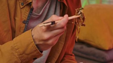Evde Asya fast food 'unun tadını çıkarıyorum. Genç adam akşam evde kanepede oturuyor, lezzetli Asya yemeği siparişi veriyor, kutusunu tutuyor, kızarmış pirinç şehriyesini çubuklarla karıştırıyor ve yiyor. Yavaş çekim