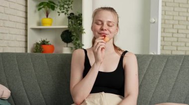 Sağlıklı gıda reklamı konsepti. Evdeki kanepede oturan ve taze vitamin meyvesi yiyen olgun şeftali yiyen, çiğneyen ve gülümseyen, atıştırmanın tadını çıkaran genç bir kadın. Yavaş çekim.