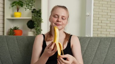 Sağlıklı gıda reklamı konsepti. Evdeki kanepede oturan genç bir kadın taze vitamin meyvesi yiyor, soyuluyor ve olgun bir muzu ısırıyor, çiğniyor ve gülümsüyor, atıştırmanın tadını çıkarıyor. Yavaş çekim.