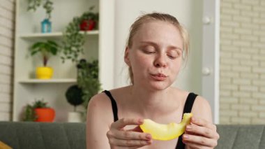 Sağlıklı gıda reklamı konsepti. Evdeki kanepede oturan ve taze vitamin meyvesi yiyen olgun kavun dilimlerini yiyen, çiğneyen ve gülümseyen, atıştırmanın tadını çıkaran genç bir kadın. Yavaş çekim.