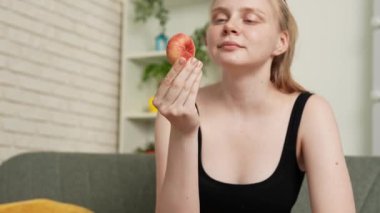 Sağlıklı gıda reklamı konsepti. Evdeki kanepede oturan ve taze vitamin meyvesi yiyen olgun şeftali yiyen, çiğneyen ve gülümseyen, atıştırmanın tadını çıkaran genç bir kadın. Yavaş çekim.