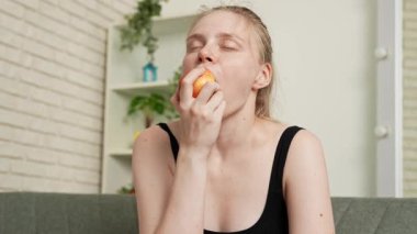 Sağlıklı gıda reklamı konsepti. Evdeki kanepede oturan ve taze vitamin meyvesi yiyen olgun elmaları yiyen, çiğneyen ve gülümseyen, atıştırmanın tadını çıkaran genç bir kadın. Yavaş çekim.