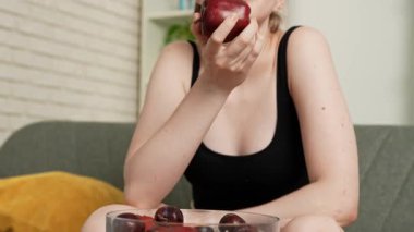 Sağlıklı gıda reklamı konsepti. Evdeki kanepede oturan ve taze vitamin meyvesi yiyen olgun kırmızı elmayı yiyen, çiğneyen ve gülümseyen, atıştırmanın tadını çıkaran genç bir kadın. Yavaş çekim.