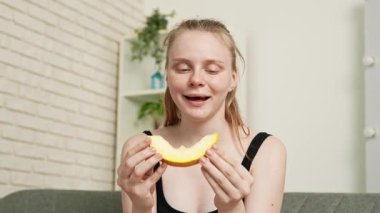Sağlıklı gıda reklamı konsepti. Evdeki kanepede oturan ve taze vitamin meyvesi yiyen olgun kavun dilimlerini yiyen, çiğneyen ve gülümseyen, atıştırmanın tadını çıkaran genç bir kadın. Yavaş çekim.