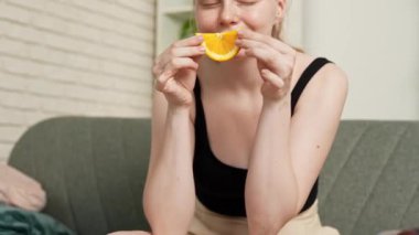 Sağlıklı gıda reklamı konsepti. Evdeki kanepede oturan ve taze vitamin meyvesi yiyen olgun portakal dilimlerini yiyen, çiğneyen ve gülümseyen, atıştırmanın tadını çıkaran genç bir kadın. Yavaş çekim.