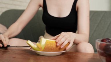 Sağlıklı gıda reklamı konsepti. Evdeki kanepede oturan ve taze vitamin meyvesi yiyen, bıçakla kesen olgun kavun dilimleri yiyen, gülümseyen, atıştırmanın tadını çıkaran genç bir kadın. Yavaş çekim.