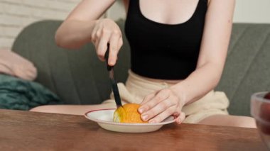 Sağlıklı gıda reklamı konsepti. Evdeki kanepede oturan ve taze vitamin meyvesi yiyen, bıçakla kesen olgun portakal dilimlerini yiyen, gülümseyen, atıştırmanın tadını çıkaran genç bir kadın. Yavaş çekim.