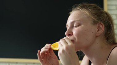 Sağlıklı gıda reklamı konsepti. Genç bir kadın evde oturup taze vitamin meyvesi yiyor olgun portakal dilimlerini yiyor, çiğniyor ve gülümsüyor, atıştırmalıkların tadını çıkarıyor, kapatıyoruz. Yavaş çekim.