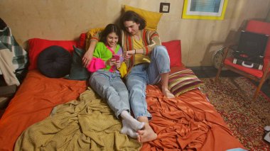 Neşeli bir çift bir yatakta oturur, bir anı paylaşır ve etrafı renkli dekorlarla çevrili akıllı bir telefona bakar..