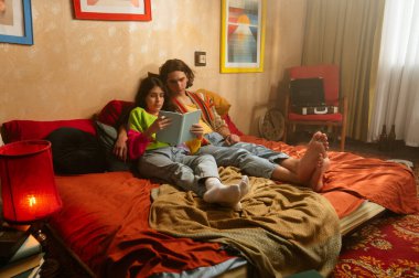 Bir çift, yatak odalarının sıcak, retro atmosferinin ortasında, bir kitaba dalmış, rahat bir şekilde otururlar..