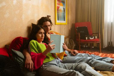 Bir çift, yatak odalarının sıcak, retro atmosferinin ortasında, bir kitaba dalmış, rahat bir şekilde otururlar..