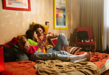 Neşeli bir çift bir yatakta oturur, bir anı paylaşır ve etrafı renkli dekorlarla çevrili akıllı bir telefona bakar..