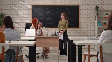 Öğrencilerin ve bir öğretmenin aktif olarak derse katıldığı bir sınıf sahnesi. Tahtada dil bilgisi noktaları gösteriliyor.