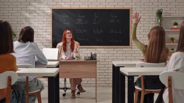 Öğrencilerin ve bir öğretmenin aktif olarak derse katıldığı bir sınıf sahnesi. Tahtada dil bilgisi noktaları gösteriliyor.