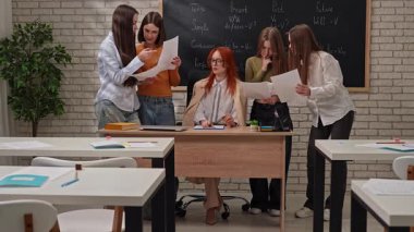 Beş öğrenci toplanıp ödevlerini gösterirken bir öğretmen masada oturuyor..