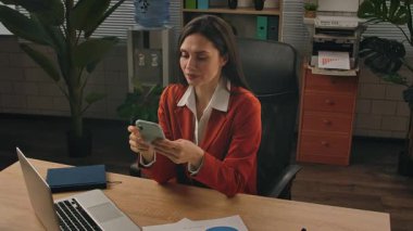 Günlük yaşam tarzı konsepti. Yaşlı yetişkin iş kadını, bitkilerle yeşil ofiste çalışıyor. Masasında oturan, dizüstü bilgisayarda çalışan, akıllı telefondan sosyal medyaya kayan bir kadın..