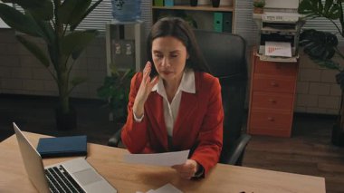 Günlük yaşam tarzı konsepti. Yaşlı, yetişkin iş kadını, bitkilerle yeşil ofiste çalışıyor. Masasında oturan, dizüstü bilgisayarda çalışan, kağıt belgelerini inceleyen, hatalar bulan, hayal kırıklığına uğramış deli ifadesi takınan bir kadın..