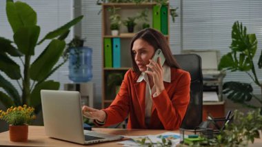 Günlük yaşam tarzı konsepti. Yaşlı, yetişkin iş kadını, bitkilerle yeşil ofiste çalışıyor. Masasında oturan, dizüstü bilgisayarda çalışan, klavyede yazan ve akıllı telefondan konuşan bir kadın..