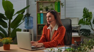 Günlük yaşam tarzı konsepti. Yaşlı yetişkin iş kadını yeşil ofiste bitkilerle çalışıyor. Masasında oturan, dizüstü bilgisayarda çalışan, klavyede yazan ve kulaklıkla konuşan bir kadın..