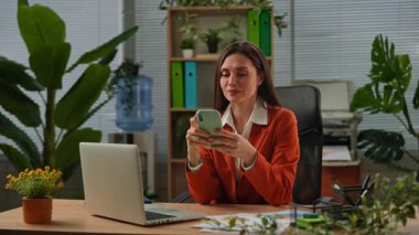 Günlük yaşam tarzı konsepti. Yaşlı yetişkin iş kadını, bitkilerle yeşil ofiste çalışıyor. Masasında oturan, dizüstü bilgisayarda çalışan, akıllı telefondan sosyal medyaya kayan bir kadın..