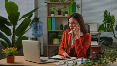 Günlük yaşam tarzı konsepti. Yaşlı yetişkin iş kadını, bitkilerle yeşil ofiste çalışıyor. Masasında oturan, dizüstü bilgisayarda çalışan, akıllı telefondan konuşan ve defterde yazan bir kadın..