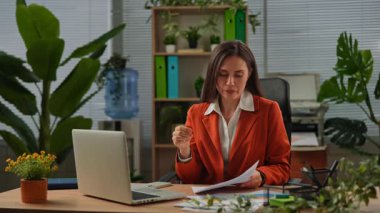 Günlük yaşam tarzı konsepti. Yaşlı yetişkin iş kadını, bitkilerle yeşil ofiste çalışıyor. Masasında oturan, dizüstü bilgisayarda çalışan, kağıt belgelerini kontrol eden, notlar yazan, bilgi okuyan bir kadın..