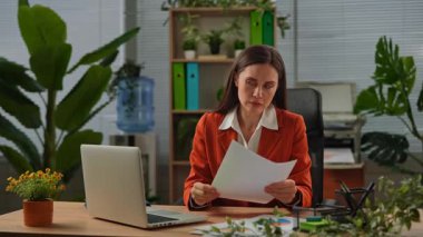 Günlük yaşam tarzı konsepti. Yaşlı, yetişkin iş kadını, bitkilerle yeşil ofiste çalışıyor. Masasında oturan, dizüstü bilgisayarda çalışan, kağıt belgelerini inceleyen, hatalar bulan, hayal kırıklığına uğramış deli ifadesi takınan bir kadın..