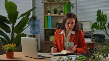 Günlük yaşam tarzı konsepti. Yaşlı, yetişkin iş kadını, bitkilerle yeşil ofiste çalışıyor. Masasında oturan, dizüstü bilgisayarda çalışan, akıllı telefondan not defteri bilgisi yazan bir kadın..