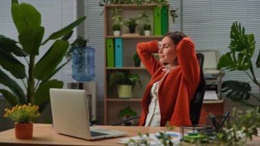 Günlük yaşam tarzı konsepti. Yetişkin bir iş kadını ofiste çalışıyor. Masasında oturan, dizüstü bilgisayarda çalışan, elleri başının arkasında oturan, gülümseyen bir kadın..