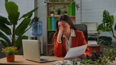 Günlük yaşam tarzı konsepti. Yaşlı yetişkin iş kadını, bitkilerle yeşil ofiste çalışıyor. Masasında oturan, dizüstü bilgisayarda çalışan, kağıt belgelerini inceleyen, hatalar bulan, çılgın ifadeler kullanan bir kadın. Yavaş çekim