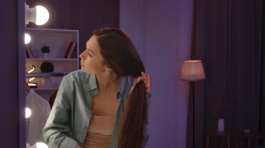 Günlük yaşam tarzı konsepti. Yaşlı yetişkin bir kadın geceyi evde, aynanın önünde uzun ipek saçlarını tarayarak, saç yaparak geçiriyor. Yavaş çekim.