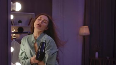 Günlük yaşam tarzı konsepti. Yetişkin bir kadın akşamını evde, aynanın önünde saç kurutma makinesiyle saç kurutarak ve şarkı söyleyerek geçirir. Yavaş çekim.