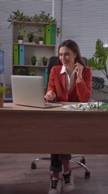 Günlük yaşam tarzı konsepti. Yaşlı, yetişkin bir iş kadını, bitkilerle yeşil ofiste çalışıyor. Masasında oturan bir kadın, harika haberler alıyor. Mutlu kazanma ifadesi. Dikey video.