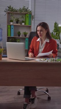 Günlük yaşam tarzı konsepti. Yaşlı yetişkin iş kadını, bitkilerle yeşil ofiste çalışıyor. Masasında oturan, dizüstü bilgisayarda çalışan, video görüşmeleri yapan, belgeleri okuyan bir kadın. Dikey video.