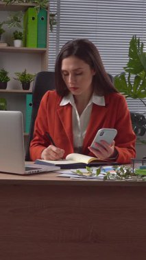 Günlük yaşam tarzı konsepti. Yaşlı yetişkin iş kadını, bitkilerle yeşil ofiste çalışıyor. Masasında oturan, dizüstü bilgisayarda çalışan, akıllı telefondan not defteri bilgisi yazan bir kadın. Dikey video.