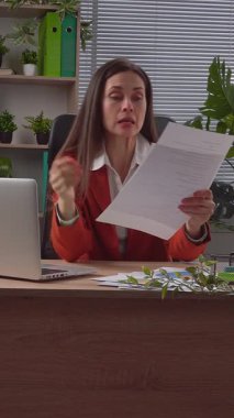 Günlük yaşam tarzı konsepti. Yaşlı yetişkin iş kadını, bitkilerle yeşil ofiste çalışıyor. Masasında oturan, dizüstü bilgisayarda çalışan, belgeleri kontrol eden, hatalar bulan, sinirli ifadeler kullanan bir kadın. Dikey video.