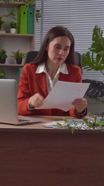 Günlük yaşam tarzı konsepti. Yaşlı yetişkin iş kadını, bitkilerle yeşil ofiste çalışıyor. Masasında oturan, dizüstü bilgisayarda çalışan, belgeleri kontrol eden, hatalar bulan, sinirli ifadeler kullanan bir kadın. Dikey video.