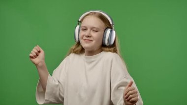Gençlik duyguları konsepti. Kameraya poz veren genç bir kız kulaklıkla müzik dinliyor, dans ediyor, pozitif ifade ediyor, krom anahtar yeşil ekran arka plan modelinde izole edilmiş. Yavaş çekim