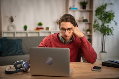 Genç bir adam, modern bir çalışma alanında dizüstü bilgisayar kullanarak evden sanal bir toplantıya katılıyor..