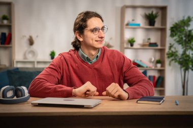 Gözlük takan ve kırmızı kazak giyen genç bir adam kendine güvenerek bir masada dizüstü bilgisayarla oturur, çalışmaya ya da çalışmaya hazırdır..