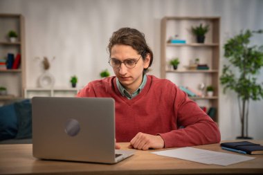 Genç bir adam, modern bir çalışma alanında dizüstü bilgisayar kullanarak evden sanal bir toplantıya katılıyor..